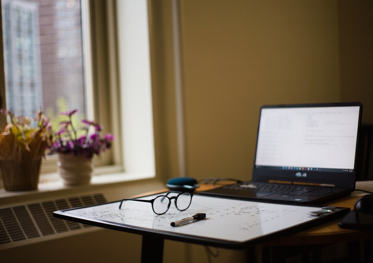 A computer on a desk with a whiteboard laying on its side in front of it and glasses on top of the whiteboard.